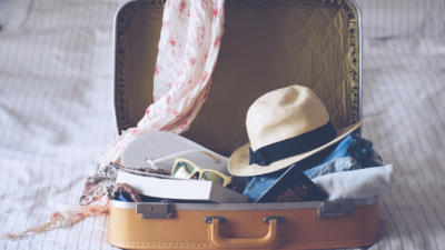 A suitcase filled with a sun hat, flip flops, passport, books, and scarf on top of a bed